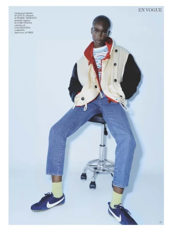 Model Maty Fall seated on a rolling stool wearing blue jeans and a color-blocked jacket for Vogue Espana.