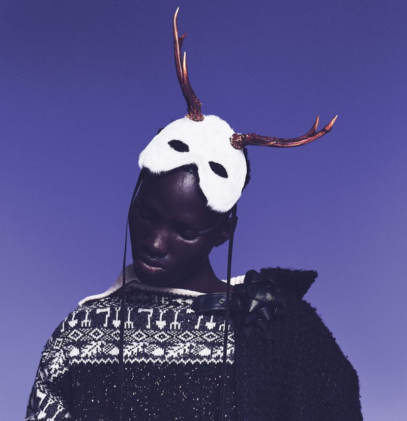 Model wearing a white animal mask with antlers and a patterned knit sweater against a solid blue background.