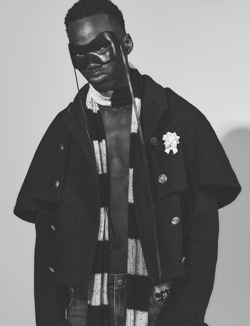 Black man wearing a leather mask and dark textured cape coat with a striped scarf against a neutral background.