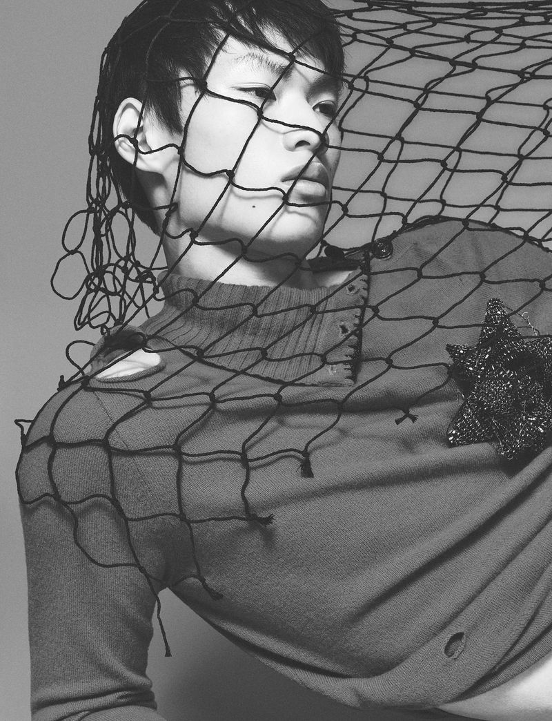 Model wearing a textured top with a net pulled over his face against a grey studio background