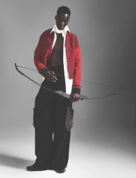 Black male model holding a bow wearing a red patterned jacket and white shirt against a neutral grey studio background