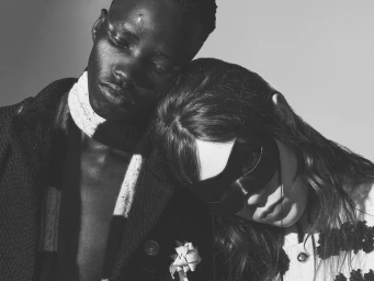 Close-up black and white portrait of two models leaning together, one wearing a dark coat and scarf and the other wearing a mask.