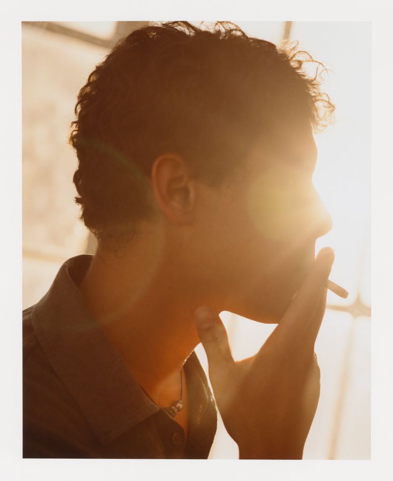 Profile of a young man smoking a cigarette in a sunlit indoor space, wearing a button-down shirt and necklace.