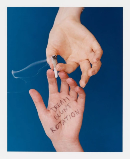 Two hands shown against a blue backdrop, one holding a smoking joint, with the words 'Dream Blunt Rotation' written on the other hand.