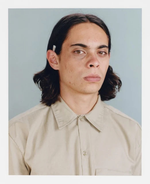 Close-up portrait of an adult male with dark hair, wearing a collared tan shirt against a light blue backdrop in a photo studio.