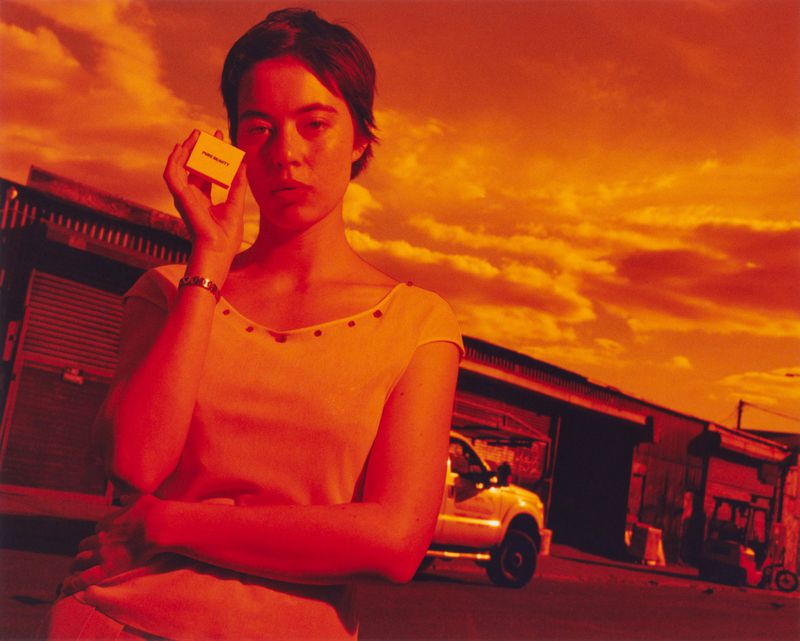 Portrait of a woman holding a small box outside with the sky and buildings in the background, all cast in red and orange light.