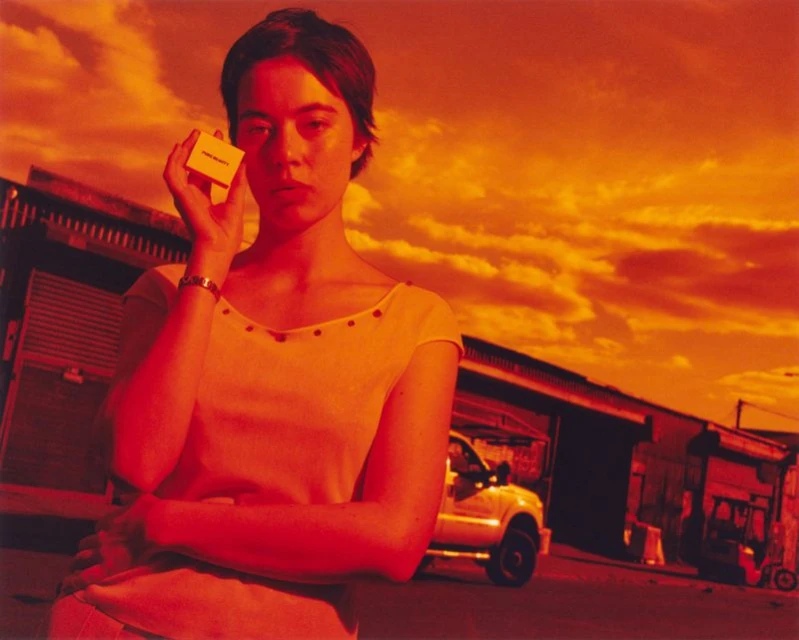 Portrait of a woman holding a small box outside with the sky and buildings in the background, all cast in red and orange light.