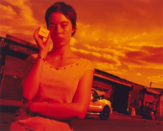 Portrait of a woman holding a small box outside with the sky and buildings in the background, all cast in red and orange light.