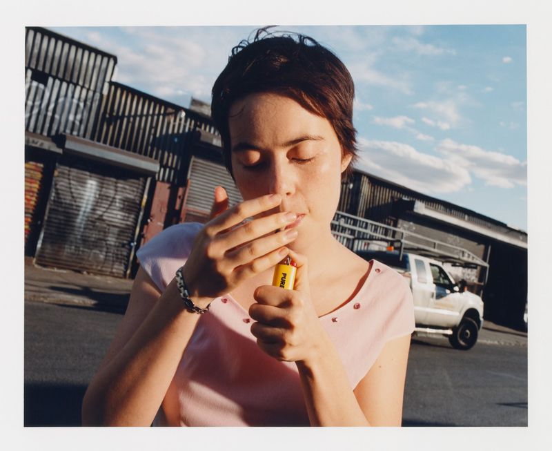 Woman in pink top lights cigarette with yellow lighter outdoors against metal storefronts and cloudy blue sky in medium shot.