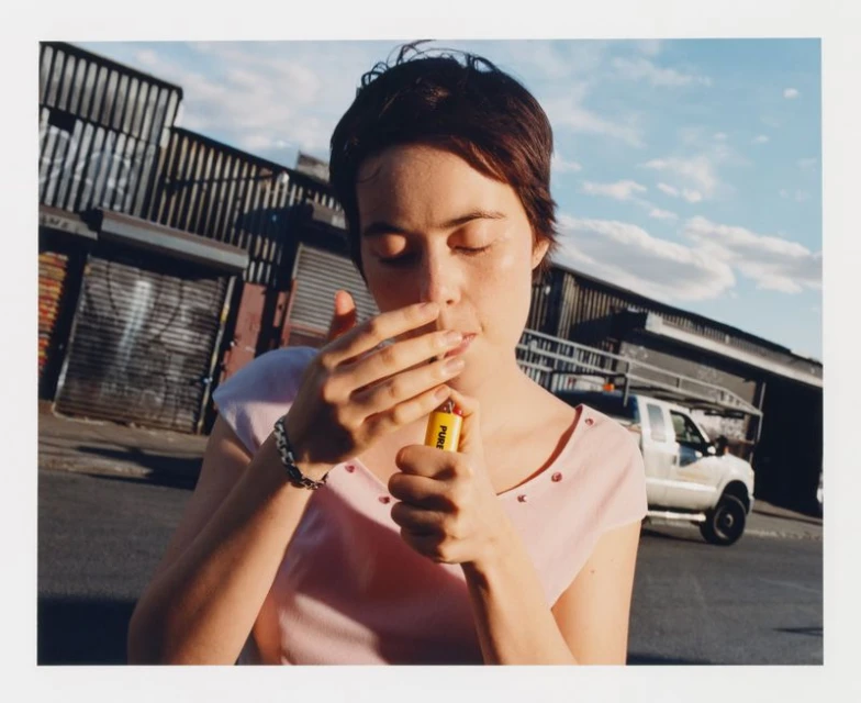Woman in pink top lights cigarette with yellow lighter outdoors against metal storefronts and cloudy blue sky in medium shot.