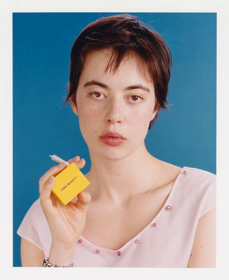 Portrait of a young woman with short dark hair, holding a yellow box and a cigarette against a blue background in a studio setting.