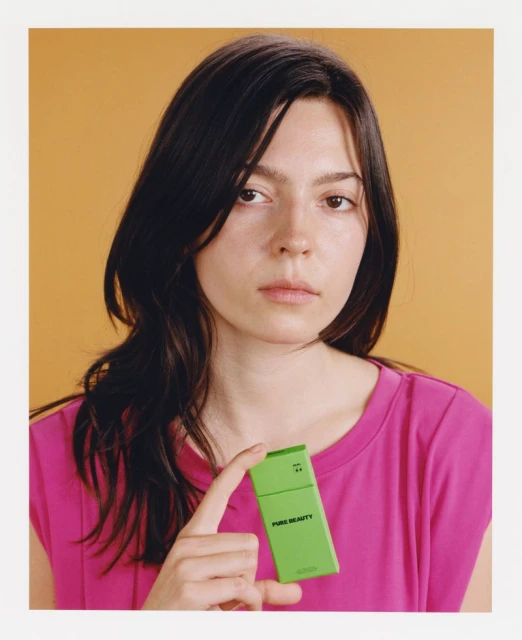 Portrait of a young brunette woman in a pink t-shirt holding a green 'Pure Beauty' product box against a solid yellow background in a studio.