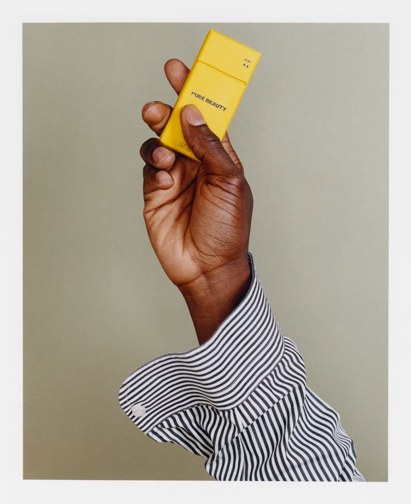 Close-up of a hand holding a yellow rectangular object with 'PURE BEAUTY' text against a light olive green background, studio lighting.