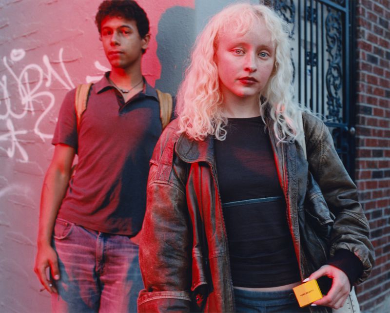 Two young adults pose outdoors by a graffiti wall; the female wears a leather jacket, and the male wears a backpack, both lit by red light.