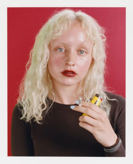 Close-up portrait of a young, light-skinned woman with wavy blonde hair and dark lipstick, holding a cigarette and yellow lighter in a studio.