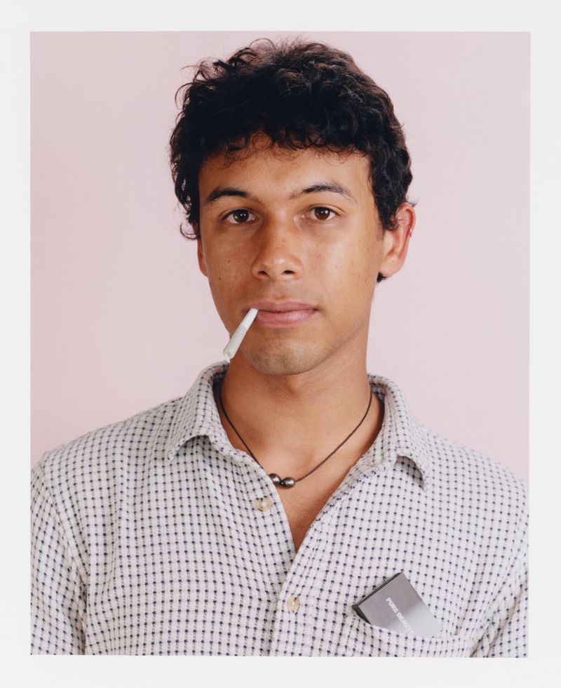 Close-up portrait of a young adult male with dark curly hair and wearing a button-down shirt, posing indoors with a light pink background.