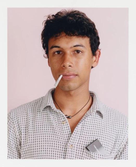 Close-up portrait of a young adult male with dark curly hair and wearing a button-down shirt, posing indoors with a light pink background.