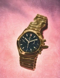 Close-up product shot of a metallic gold chronograph watch with a black dial set against a textured pink background.