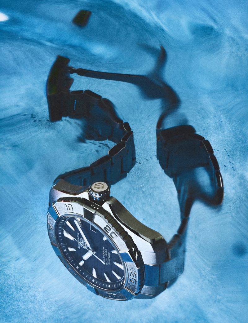 Close-up underwater shot of a stainless steel sports watch with a blue dial submerged in swirling blue water.