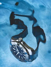 Close-up underwater shot of a stainless steel sports watch with a blue dial submerged in swirling blue water.