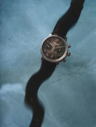 Close-up of a luxury wristwatch with a black leather strap against a mottled blue textured background.