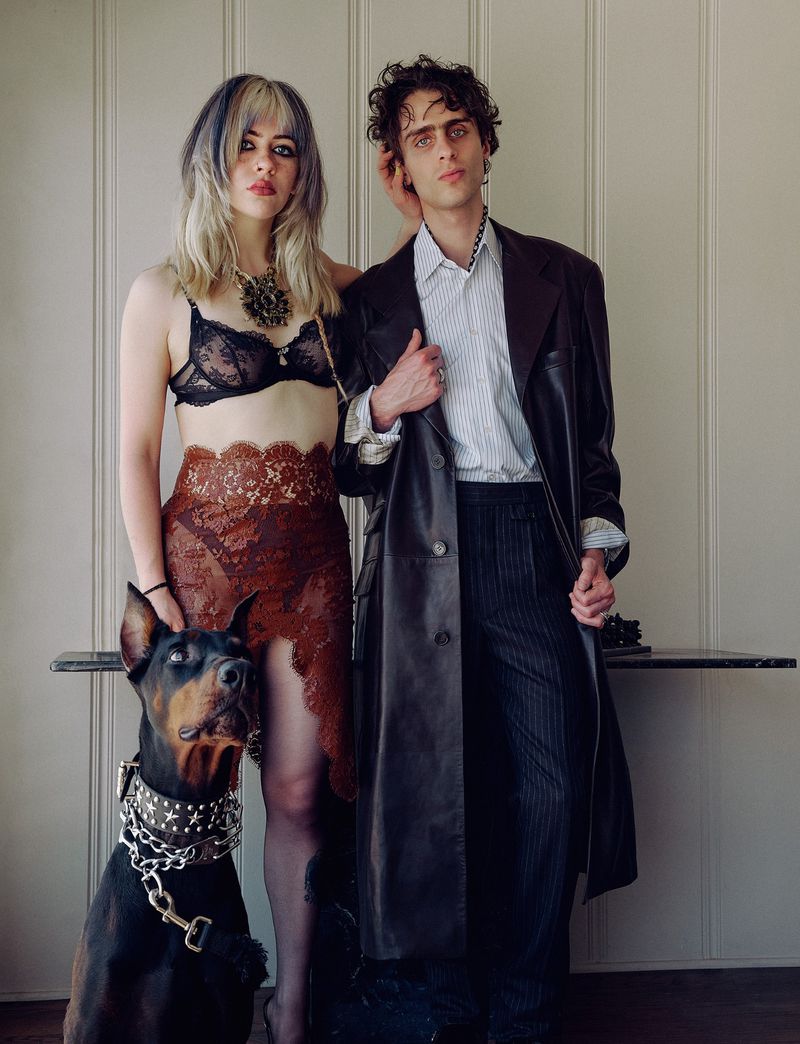 Portrait of a woman in a black lace bra and brown lace skirt standing next to a man in a black leather coat and a Doberman dog
