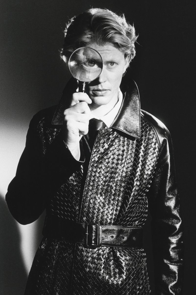 Man holding a magnifying glass over his eye while wearing a dark woven leather trench coat for HERO Magazine.