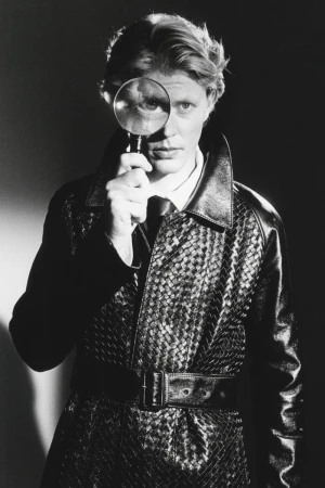 Man holding a magnifying glass over his eye while wearing a dark woven leather trench coat for HERO Magazine.