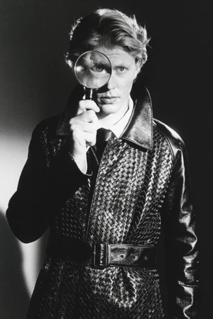 Man holding a magnifying glass over his eye while wearing a dark woven leather trench coat for HERO Magazine.