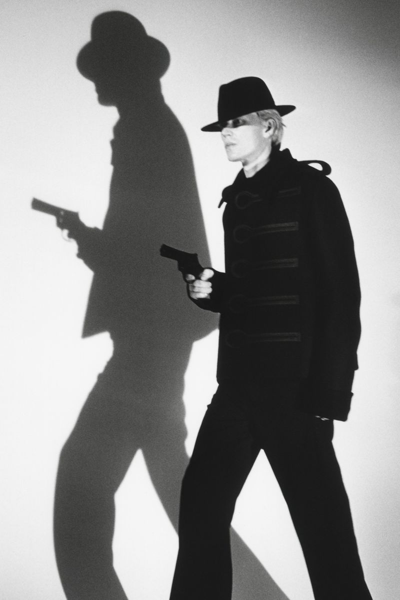 Model in black fedora holding a handgun against a white backdrop with a dramatic large shadow casting a silhouette holding a gun