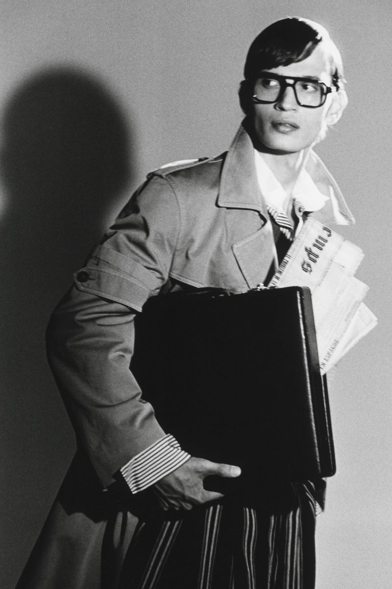Man wearing heavy-rimmed glasses and trench coat carrying a briefcase and newspapers against a plain studio background.
