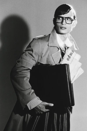 Man wearing heavy-rimmed glasses and trench coat carrying a briefcase and newspapers against a plain studio background.