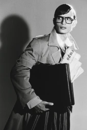 Man wearing heavy-rimmed glasses and trench coat carrying a briefcase and newspapers against a plain studio background.