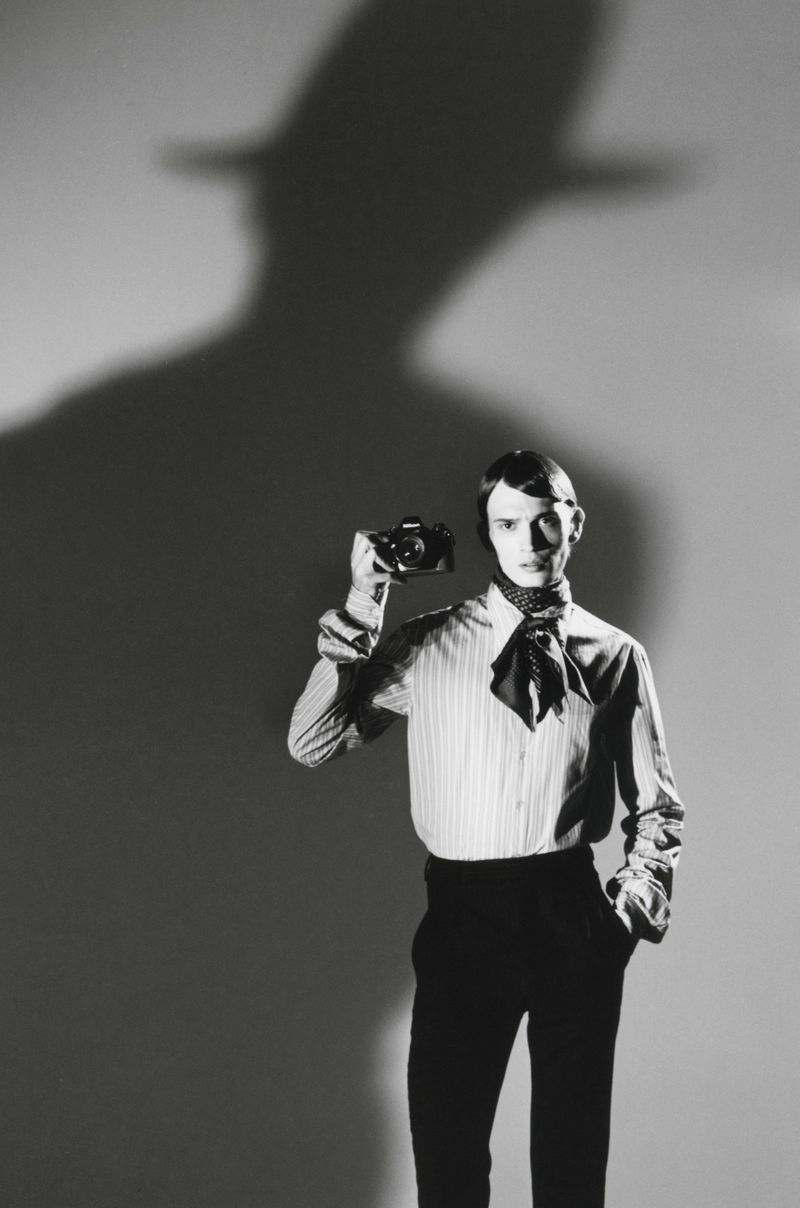 Model wearing a striped shirt and neck scarf, holding a vintage camera against a light background with a dramatic shadow above.
