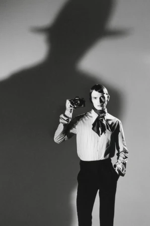 Model wearing a striped shirt and neck scarf, holding a vintage camera against a light background with a dramatic shadow above.