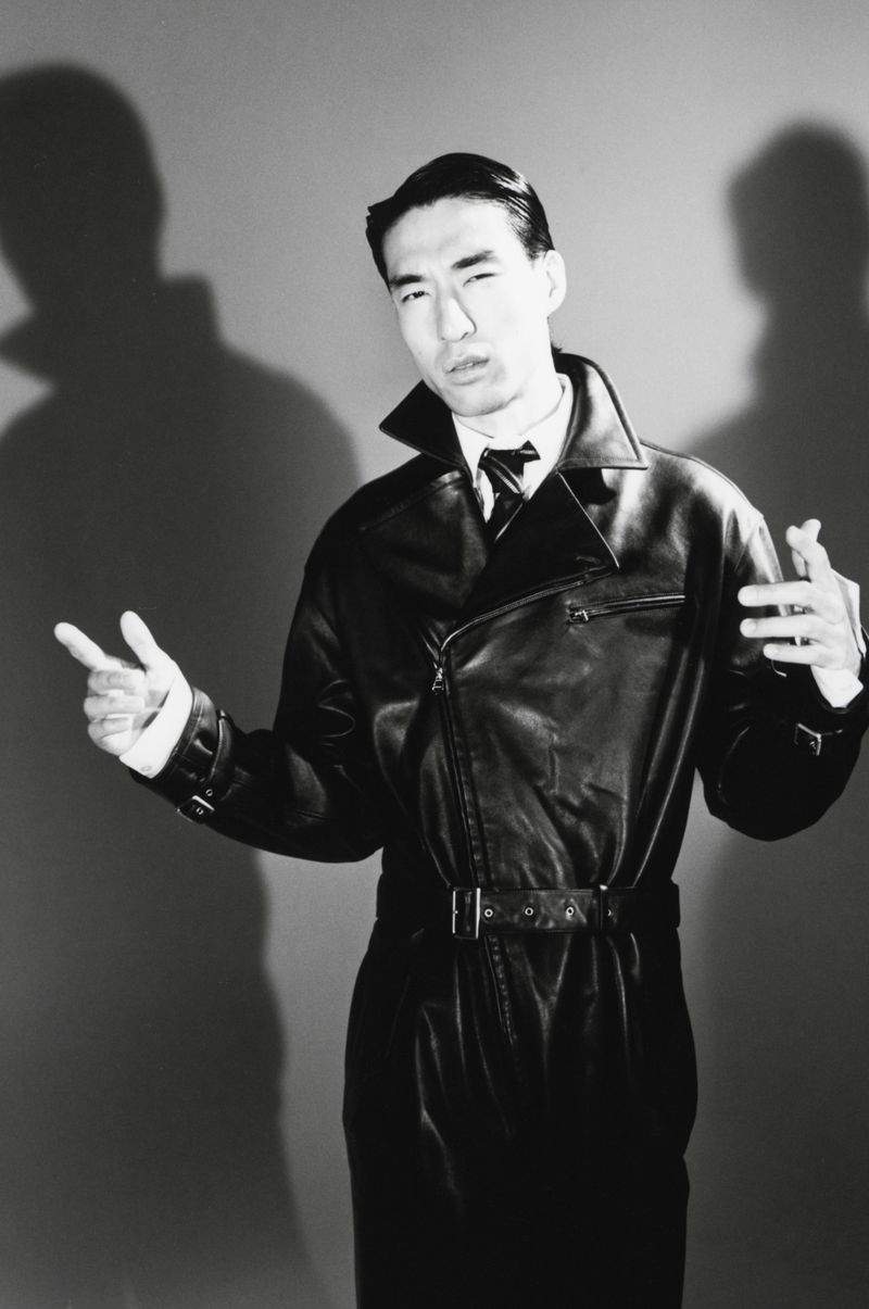 Man wearing a black leather trench coat posing against a grey seamless studio backdrop with dramatic shadows