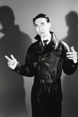 Man wearing a black leather trench coat posing against a grey seamless studio backdrop with dramatic shadows