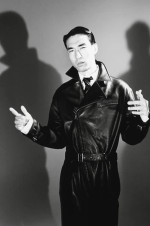 Man wearing a black leather trench coat posing against a grey seamless studio backdrop with dramatic shadows
