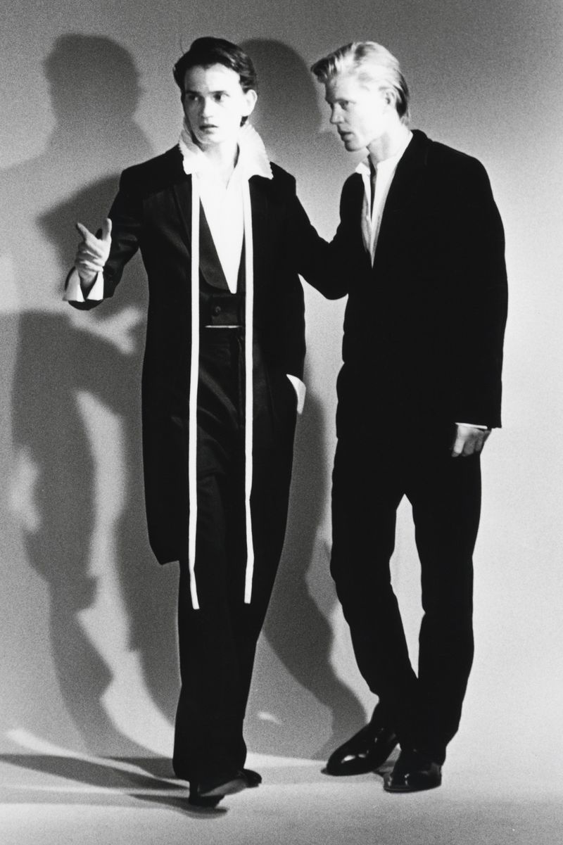Two male models in black suits and white shirts posing against a grey studio backdrop with strong shadows