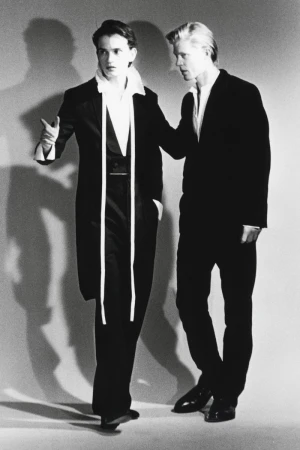 Two male models in black suits and white shirts posing against a grey studio backdrop with strong shadows