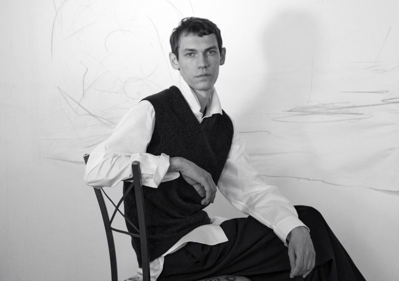 Black and white portrait of a male model seated backward on a chair, wearing a white shirt and dark sweater vest.