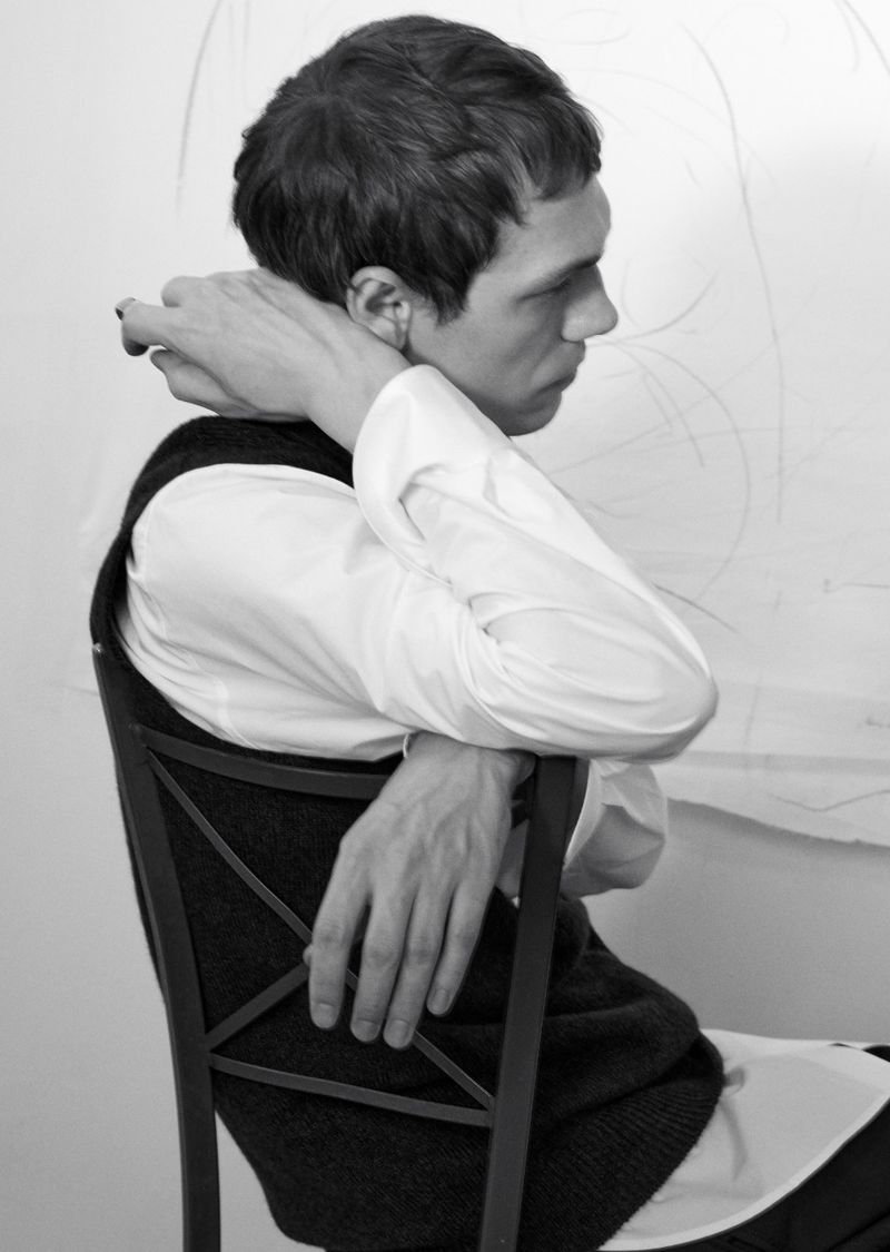 Profile view of male model seated on wooden chair wearing white shirt and dark vest against a plain light wall.