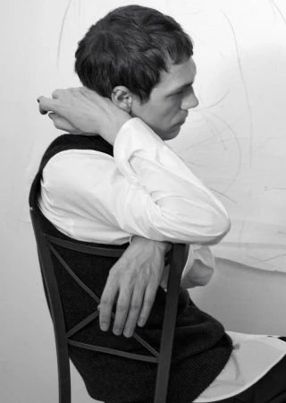 Profile view of male model seated on wooden chair wearing white shirt and dark vest against a plain light wall.