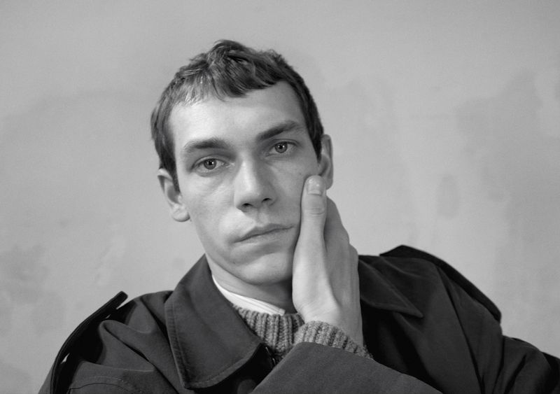Portrait of a male model with his hand to his cheek wearing a dark coat and textured sweater against a plain background.