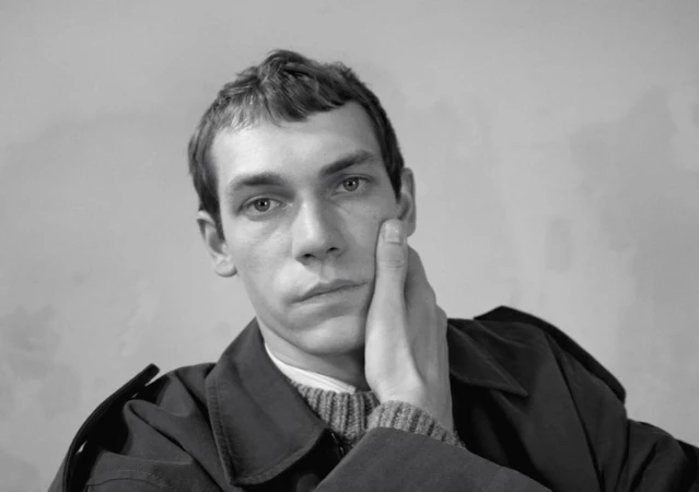 Portrait of a male model with his hand to his cheek wearing a dark coat and textured sweater against a plain background.