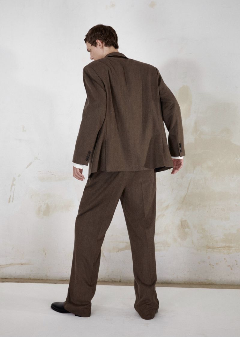 Model wearing a brown suit seen from behind against a textured light wall and white floor for Harper's Bazaar France Homme.