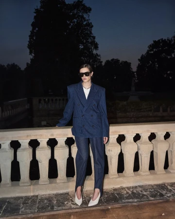 Model Kim Schell wearing a blue pinstripe blazer and stirrup pants leaning on an outdoor stone balustrade at dusk