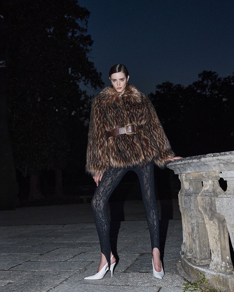 Model wearing a fuzzy brown fur jacket and black lace leggings outdoors at night by a stone balustrade