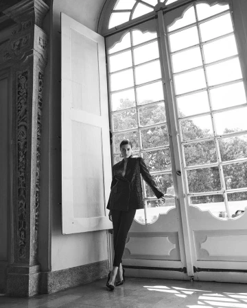 Model Kim Schell leaning against a tall window wearing a dark blazer and trousers in AMICA Magazine editorial by David Roemer