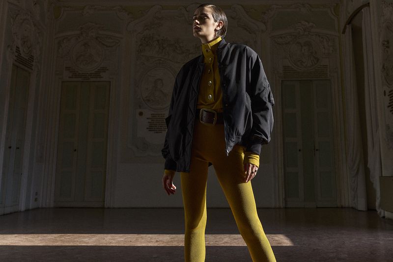 Model wearing a black bomber jacket over a mustard yellow turtleneck and trousers in an ornate room.